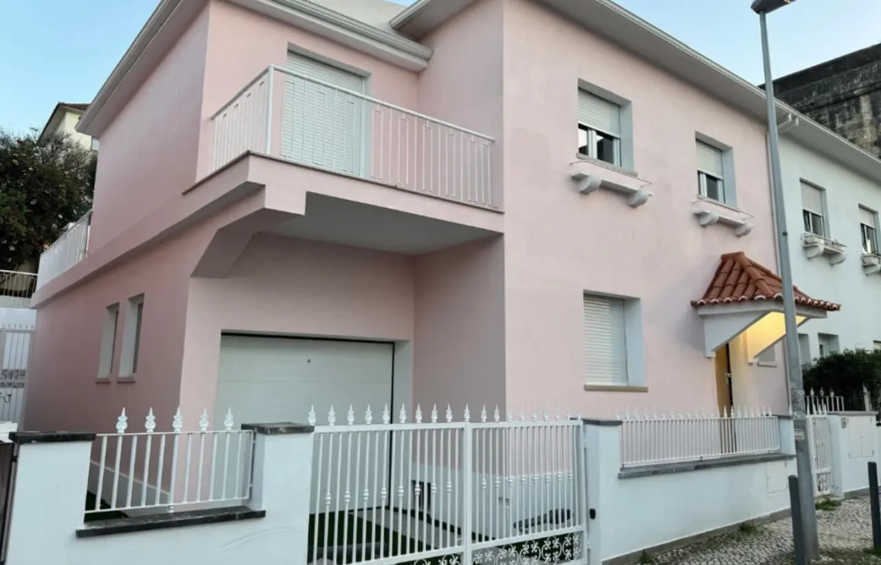 Fachada cor-de-rosa da moradia, com varanda, garagem e portão branco.