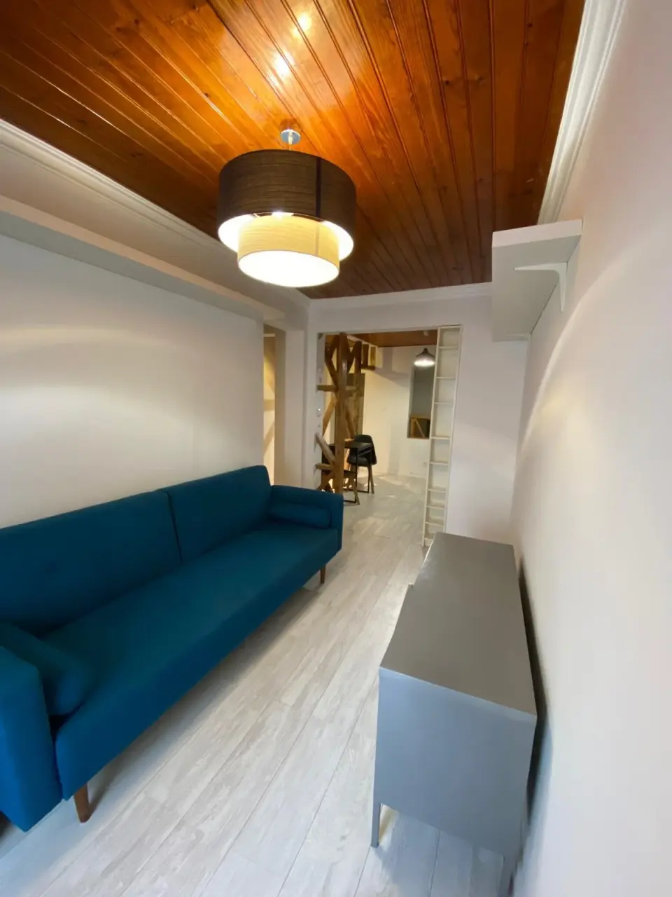 Sala com teto em madeira, sofá azul e candeeiro suspenso.