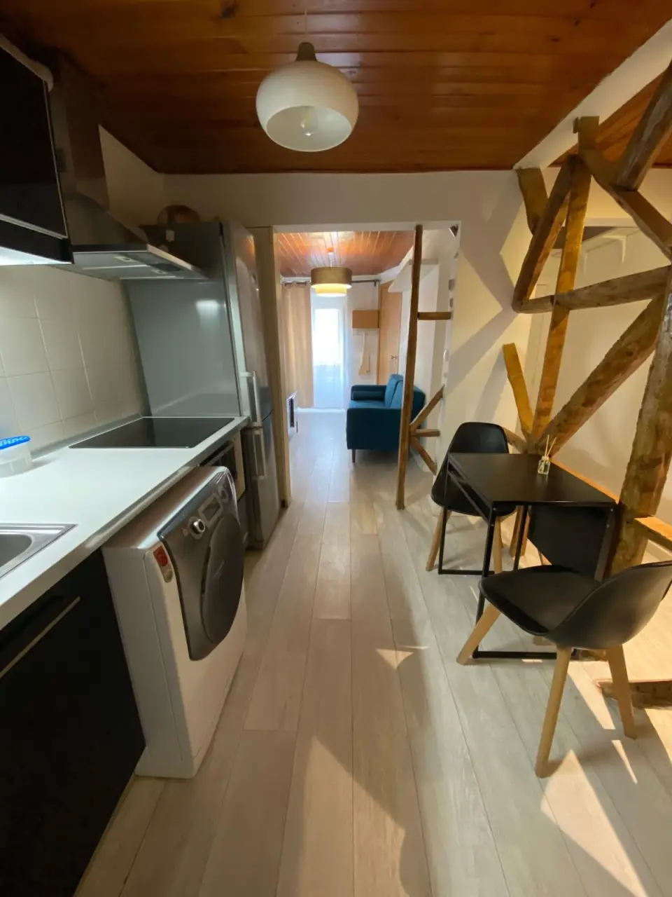 Cozinha e zona de refeições junto a vigas de madeira em cruz, com a sala ao fundo.