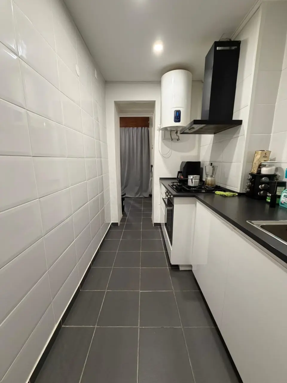 Cozinha com azulejo branco, móveis brancos, bancada preta e pavimento cinzento.